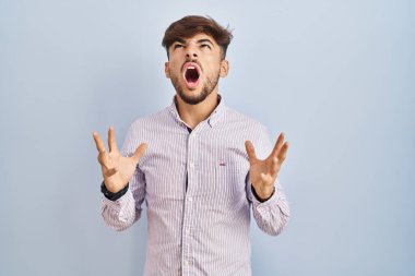 Arab man with beard standing over blue background crazy and mad shouting and yelling with aggressive expression and arms raised. frustration concept. 