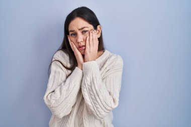 Genç esmer kadın mavi arka planda duruyor. Yorgun eller yüzünü kapatıyor, depresyon ve üzüntü, üzgün ve sorunlardan rahatsız. 