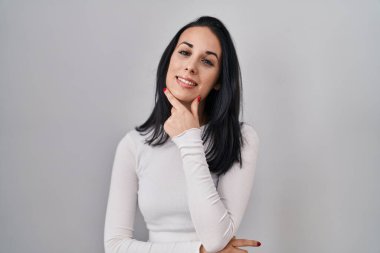 Hispanic woman standing over isolated background looking confident at the camera smiling with crossed arms and hand raised on chin. thinking positive. 