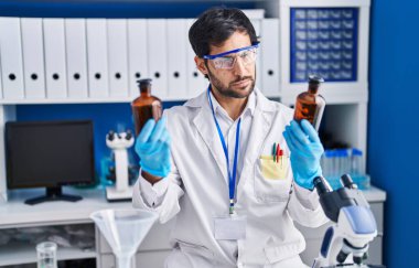 Young hispanic man scientist holding bottles at laboratory