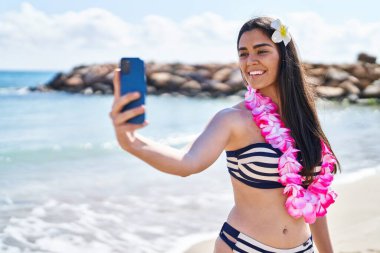 Bikini giyen genç İspanyol kadın deniz kenarında akıllı telefonla selfie çekiyor.