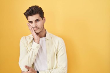 Sarı arka planda duran İspanyol genç adam çapraz kollarla depresyon sorunlarından yorgun ve sıkılmış göründüğünü düşünüyor.. 