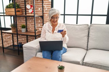 Evdeki koltukta oturan orta yaşlı bir kadın dizüstü bilgisayar ve kredi kartı kullanıyor.