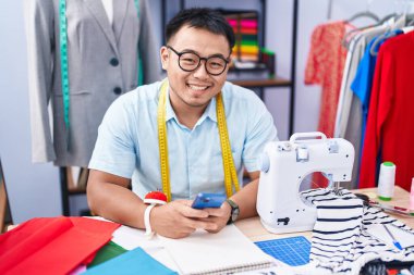 Young chinese man tailor smiling confident using smartphone at tailor shop