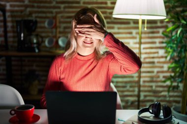 Blonde woman using laptop at night at home smiling and laughing with hand on face covering eyes for surprise. blind concept.  clipart