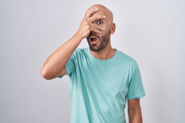 Middle age bald man standing over white background peeking in shock covering face and eyes with hand, looking through fingers with embarrassed expression. 
