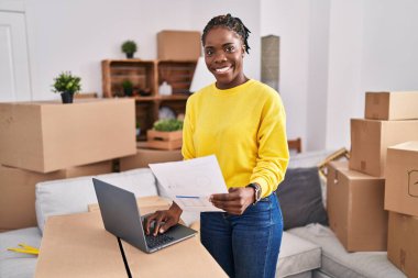 African american woman using laptop holding document at new home