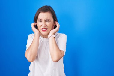 Orta yaşlı Latin bir kadın mavi arka planda duruyor. Yüksek sesli müzik sesi için parmaklarıyla kulaklarını kapatıyor. sağır kavramı. 