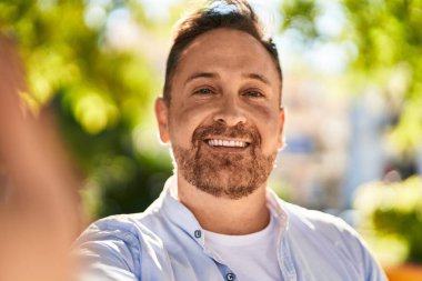 Genç, beyaz bir adam gülümsüyor. Parkta kamerayla selfie çekiyor.