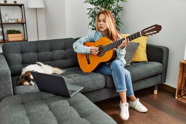 Beyaz bir kadın online klasik gitar dersi alıyor. Evde köpekle birlikte kanepede oturuyor.