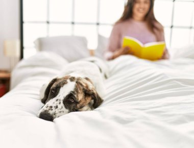 Yatak odasında köpekle kanepede kitap okuyan genç bir kadın.