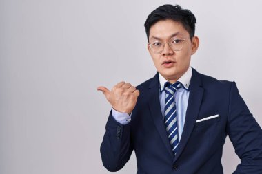 Young asian man wearing business suit and tie surprised pointing with hand finger to the side, open mouth amazed expression. 