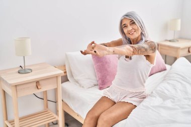 Orta yaşlı, kır saçlı bir kadın yatak odasında kollarını açarak uyanıyor.