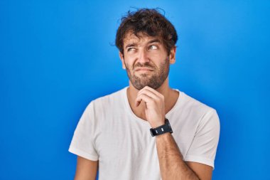 Mavi arka planda dikilen Latin kökenli genç adam bir soru hakkında endişeli ve gergin bir şekilde düşünüyor. 