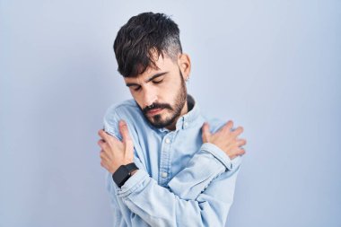 Mavi arka planda sakallı genç İspanyol adam kendine sarılıyor mutlu ve pozitif, kendinden emin gülümsüyor. Sevgi ve özgüven. 