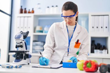 Young beautiful hispanic woman scientist weighing piece of apple writing report at laboratory