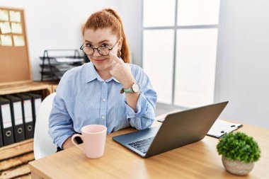 Ofiste çalışan genç kızıl saçlı bir kadın bilgisayarı kullanarak gözüne işaret ediyor jestini izliyor, şüpheli bir ifade. 