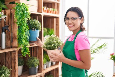 Young hispanic woman florist holding plant of shelving at florist