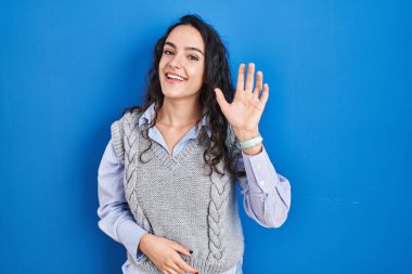 Mavi arka planda duran genç esmer kadın selam veriyor mutlu ve gülümsüyor, dostça karşılama hareketi yapıyor. 