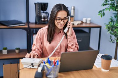 Genç İspanyol iş kadını laptopunu kullanıyor ve ofisteki akıllı telefondan konuşuyor.