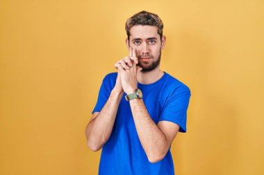 Hispanic man with beard standing over yellow background holding symbolic gun with hand gesture, playing killing shooting weapons, angry face 