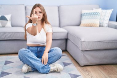 Young caucasian woman sitting on the floor at the living room showing middle finger, impolite and rude fuck off expression 