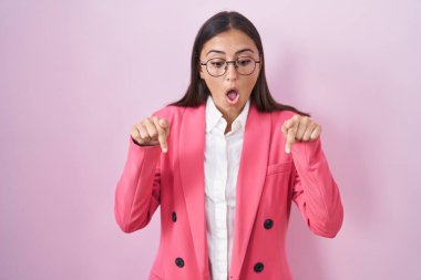 Young hispanic woman wearing business clothes and glasses pointing down with fingers showing advertisement, surprised face and open mouth 