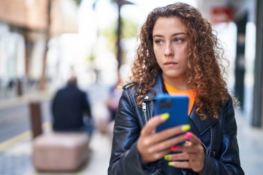 Genç, güzel, İspanyol bir kadın sokakta rahat bir ifadeyle akıllı telefon kullanıyor.