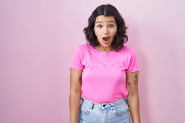 Young hispanic woman standing over pink background afraid and shocked with surprise and amazed expression, fear and excited face. 