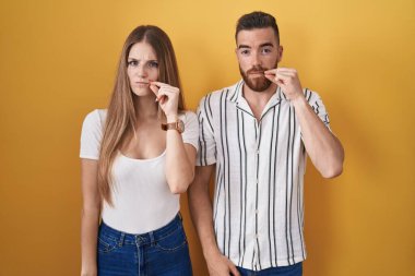 Young couple standing over yellow background mouth and lips shut as zip with fingers. secret and silent, taboo talking 