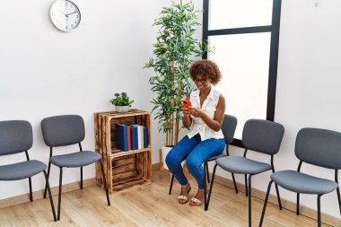 Bekleme odasında sandalyede oturan akıllı telefon kullanan Afrikalı Amerikalı genç bir kadın.