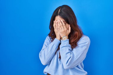 Hispanic young woman standing over blue background with sad expression covering face with hands while crying. depression concept. 