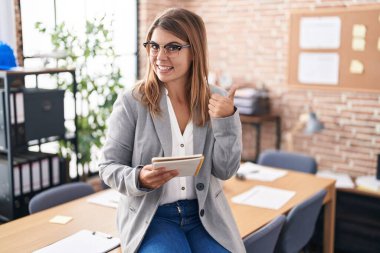 Young hispanic woman working at the office wearing glasses doing happy thumbs up gesture with hand. approving expression looking at the camera showing success. 