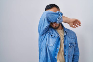 Young hispanic man standing over isolated background covering eyes with arm smiling cheerful and funny. blind concept. 