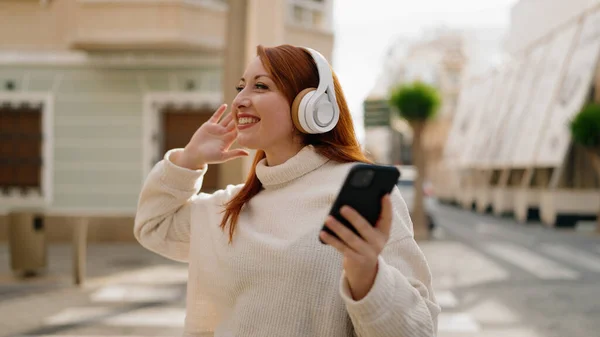 若い赤毛の女性が路上でスマートフォンを使って自信を持って音楽を聞いて笑っている — ストック写真