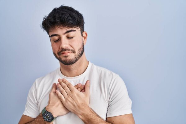 Hispanic man with beard standing over white background smiling with hands on chest with closed eyes and grateful gesture on face. health concept. 