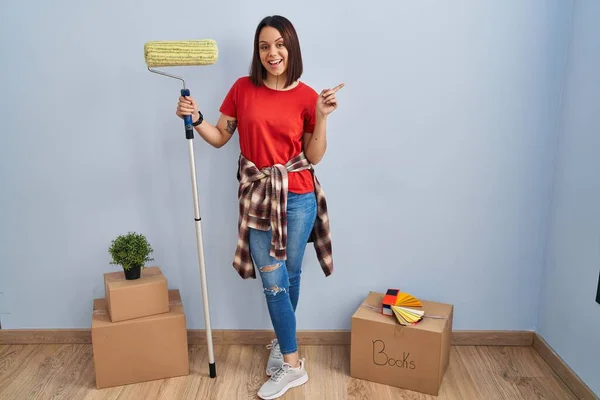 Young hispanic woman painting home walls with paint roller with a big smile on face, pointing with hand finger to the side looking at the camera.