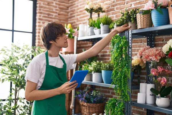 Young blond man florist using touchpad holding plant at flower shop