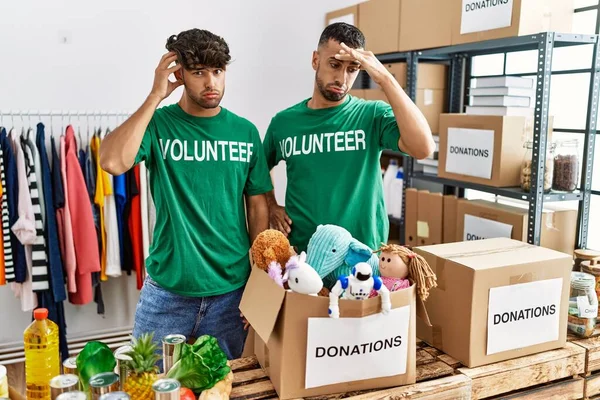 Young gay couple wearing volunteer t shirt at donations stand worried and stressed about a problem with hand on forehead, nervous and anxious for crisis