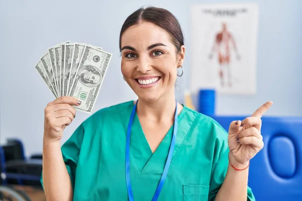年轻而惊慌的女人拿着钞票在疼痛康复诊所工作 满面笑容 手指手画脚地朝旁边看去 — 图库照片