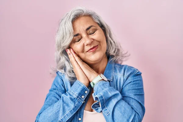 Middle age woman with grey hair standing over pink background sleeping tired dreaming and posing with hands together while smiling with closed eyes.