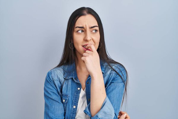 Hispanic woman standing over blue background looking stressed and nervous with hands on mouth biting nails. anxiety problem. 