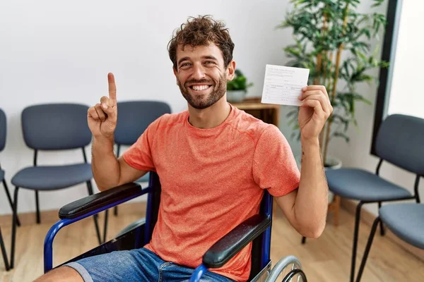 Young handsome man holding covid record card sitting on wheelchair smiling with an idea or question pointing finger with happy face, number one