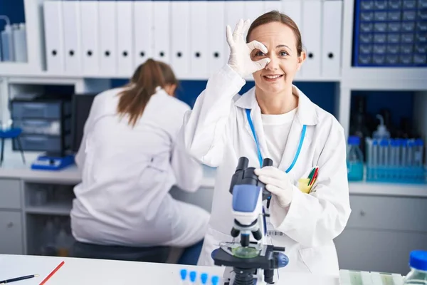 Blonde woman working on cruelty free laboratory smiling happy doing ok sign with hand on eye looking through fingers