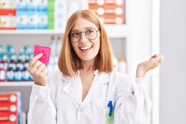 Young Redhead Woman Working Pharmacy Drugstore Holding Condom Screaming Proud — Zdjęcie stockowe