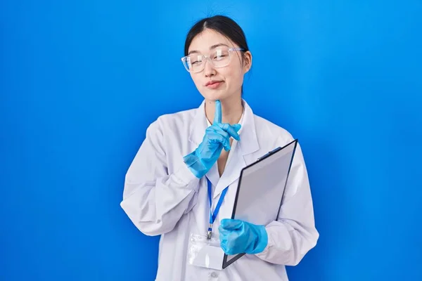 Chinese young woman working at scientist laboratory thinking concentrated about doubt with finger on chin and looking up wondering