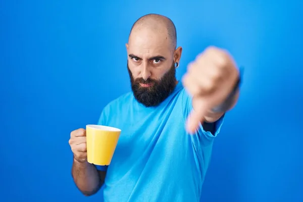 Young hispanic man with beard and tattoos drinking a cup of coffee looking unhappy and angry showing rejection and negative with thumbs down gesture. bad expression.