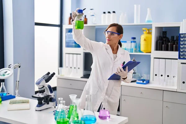 中年妇女穿着科学家制服 拿着剪贴板和瓶子在实验室里 — 图库照片