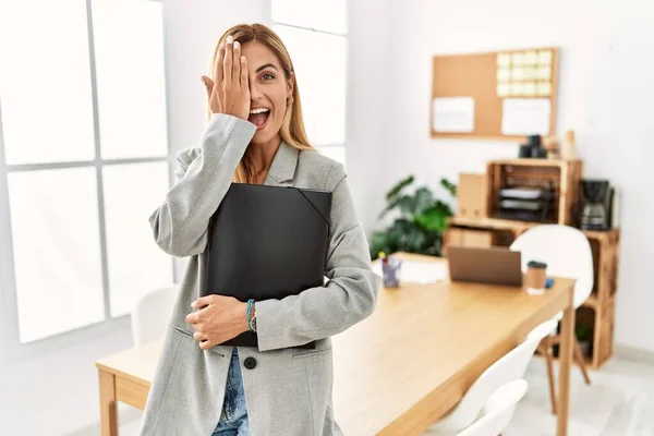 Blonde business woman at the office covering one eye with hand, confident smile on face and surprise emotion.