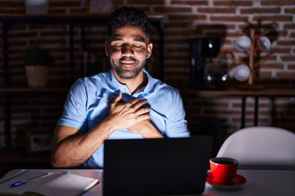 Hispanic man with beard using laptop at night smiling with hands on chest with closed eyes and grateful gesture on face. health concept.